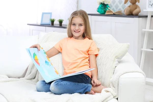 Cute little girl with book sitting on sofa at home — Stock Photo, Image