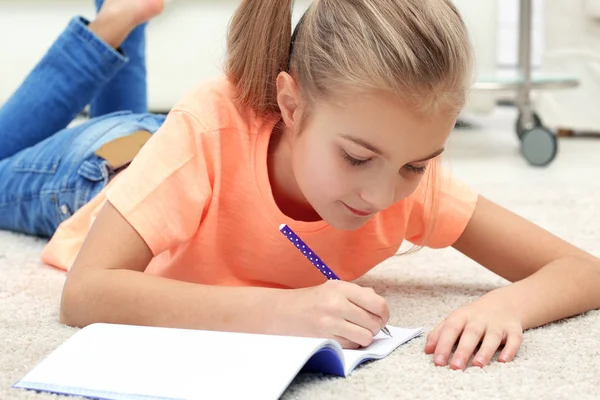 Attractive little girl lying on the floor and writing in copybook — Stock Photo, Image