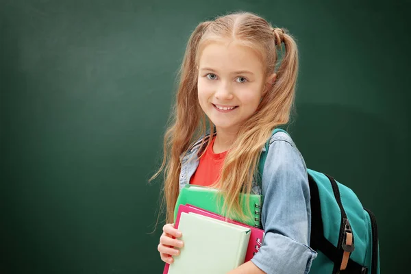 Attractive little girl with books standing on green blackboard background — Stock Photo, Image