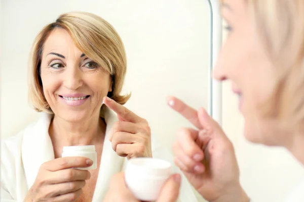 Donna anziana che applica la crema cosmetica su faccia vicino a specchio — Foto Stock