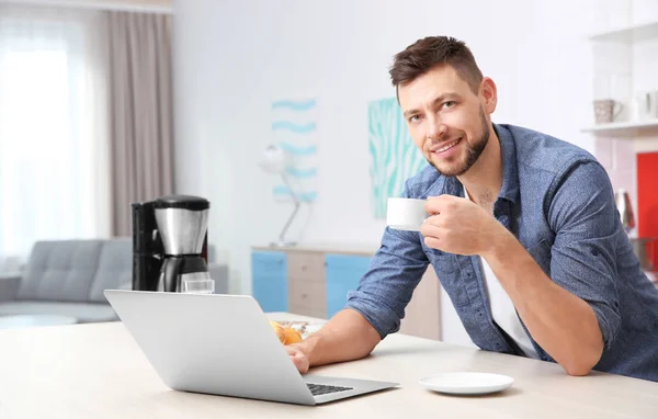 Guapo joven bebiendo café mientras trabaja con el ordenador portátil en la cocina — Foto de Stock