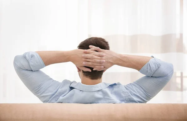 Young man resting on couch with hands behind his head in light room — Stock Photo, Image