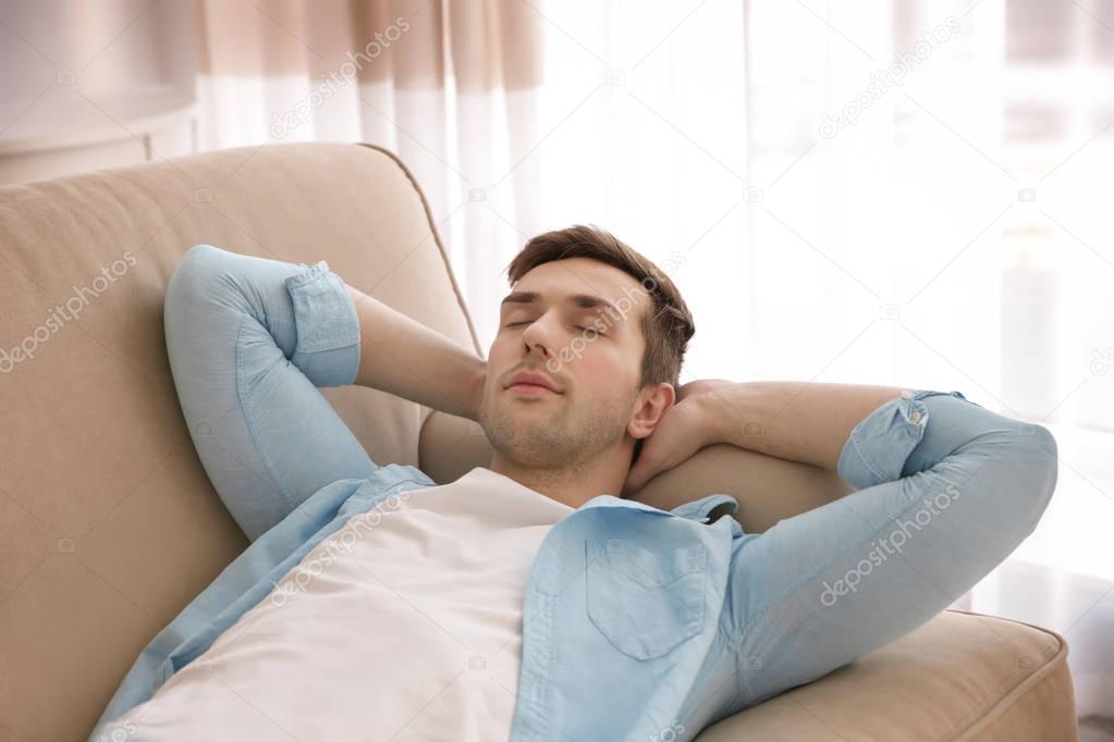 Young man resting on couch with hands behind his head in light room