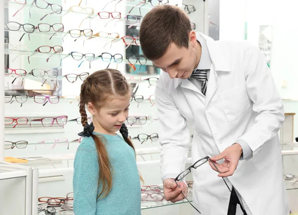 Joven médico ayudando a la niña a elegir nuevas gafas en la tienda —  Fotos de Stock