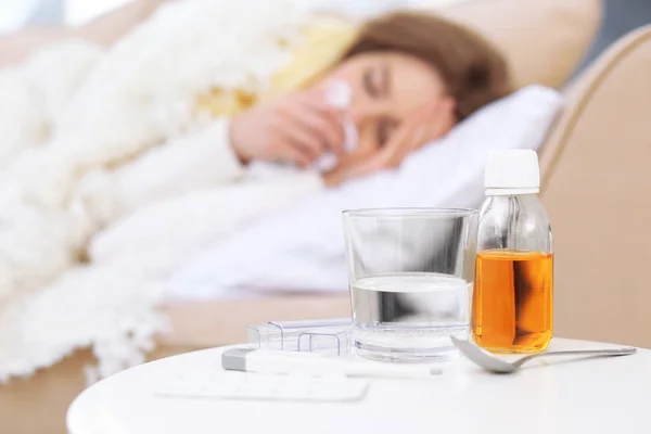 Medicamentos, termómetro electrónico y vaso de agua con la mujer enferma borrosa en el fondo —  Fotos de Stock