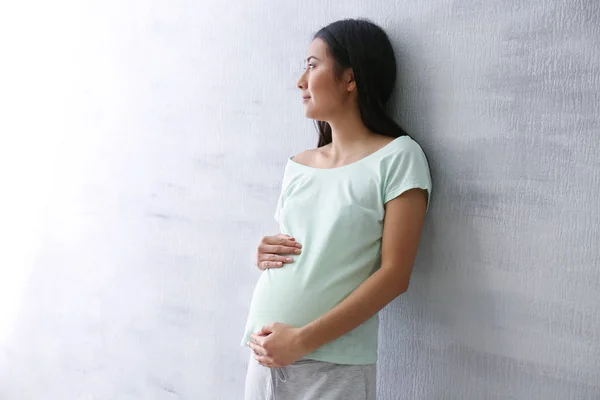 Pregnant Asian woman standing near light wall — Stock Photo, Image