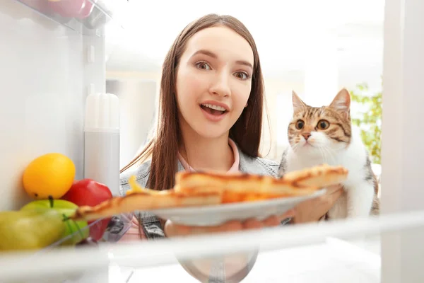Молодая женщина и симпатичная кошка собираются съесть вкусную пиццу из холодильника — стоковое фото