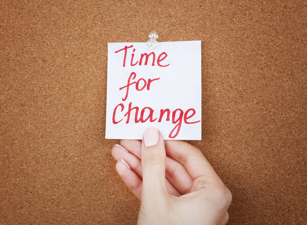 Female hand holding note with phrase TIME FOR CHANGE on cork board background