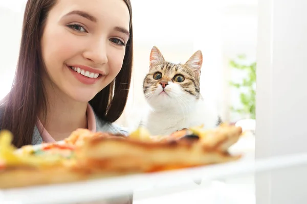 Молодая женщина и симпатичная кошка собираются съесть вкусную пиццу из холодильника — стоковое фото