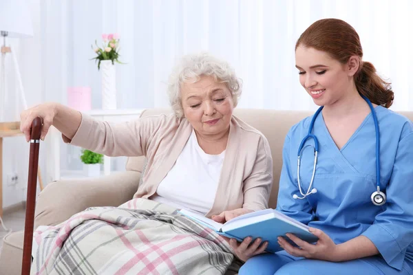 Enfermeira livro de leitura para a mulher idosa no sofá — Fotografia de Stock
