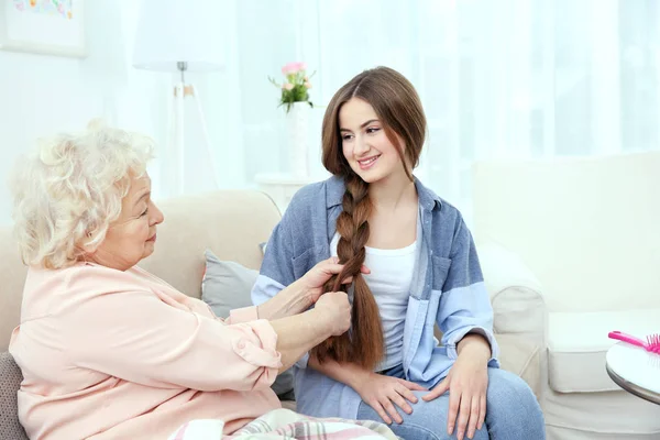 Grandmother pigtails braiding granddaughter's hair on couch at home — Stock Photo, Image