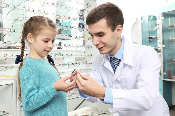 Joven médico ayudando a la niña a elegir nuevas gafas en la tienda —  Fotos de Stock