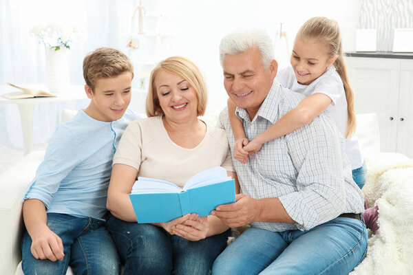 Senior couple reading book to grandchildren while sitting on sofa in living room