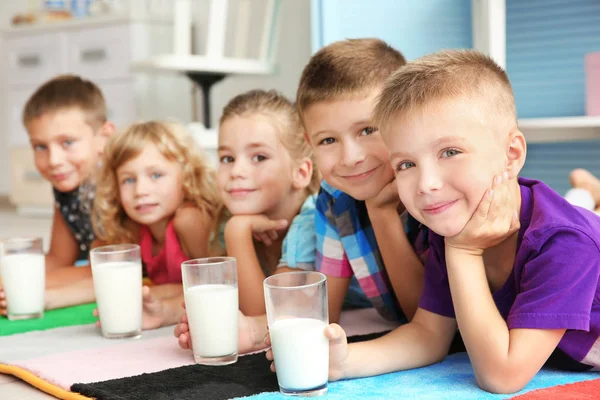 Crianças alegres com copos de leite deitado no tapete colorido na sala — Fotografia de Stock