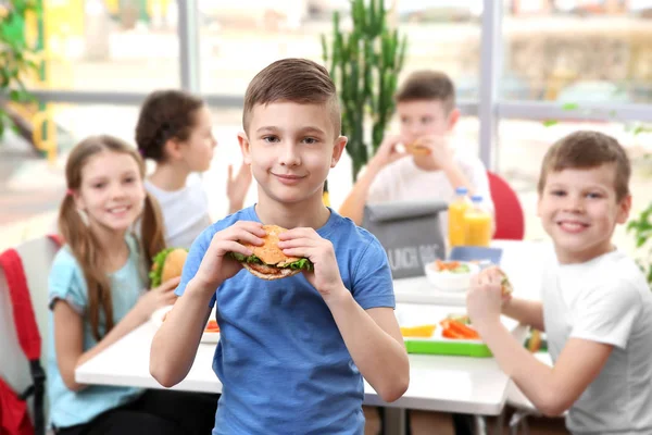 Söt Pojke Äter Hamburgare Och Barn Sitter Vid Bord Skolmatsalen — Stockfoto