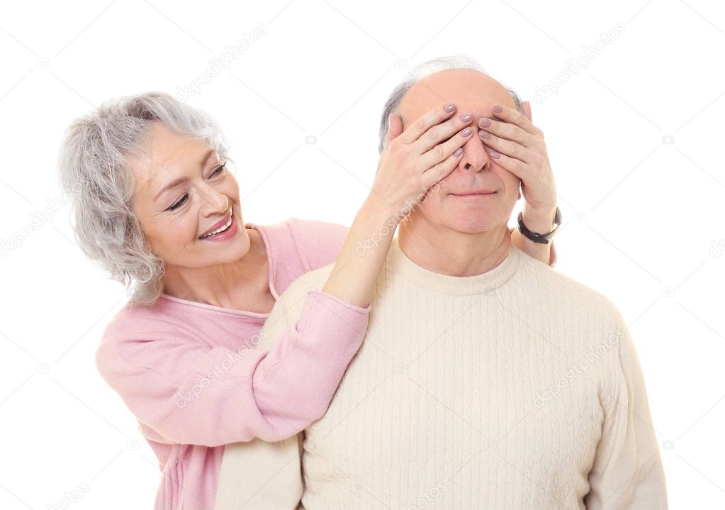 Happy senior woman covering her husband's eyes on white background