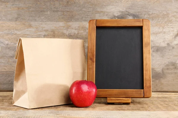 Paper lunch bag, red apple and small blackboard on wooden background — Stock Photo, Image