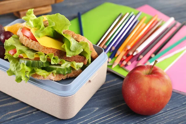 Scatola da pranzo con cibo delizioso, matite e mela su sfondo di legno blu — Foto Stock