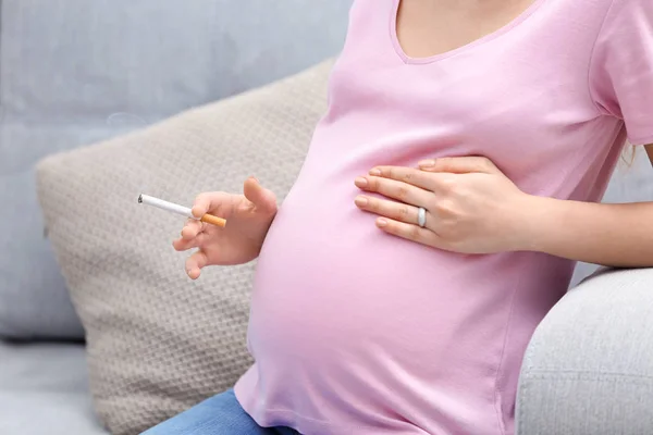 Беременная женщина курит сигарету сидя дома на диване — стоковое фото