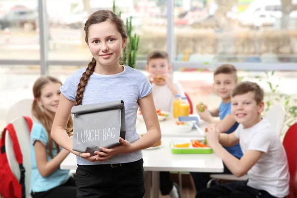 Söt flicka håller lunch-väska och barn äta vid bordet i skolmatsalen — Stockfoto