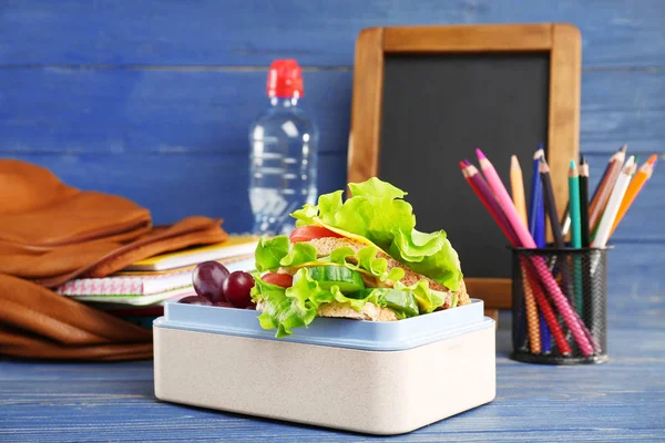 Scatola da pranzo con delizioso cibo e cancelleria su sfondo blu in legno — Foto Stock