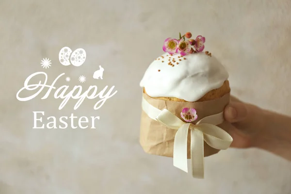 Text HAPPY EASTER and female hand holding traditional cake on light background — Stock Photo, Image