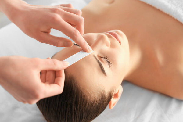 Beautician waxing young woman's eyebrows in spa center