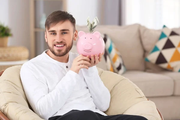 Jovem feliz sentado em poltrona com banco porquinho e dinheiro — Fotografia de Stock