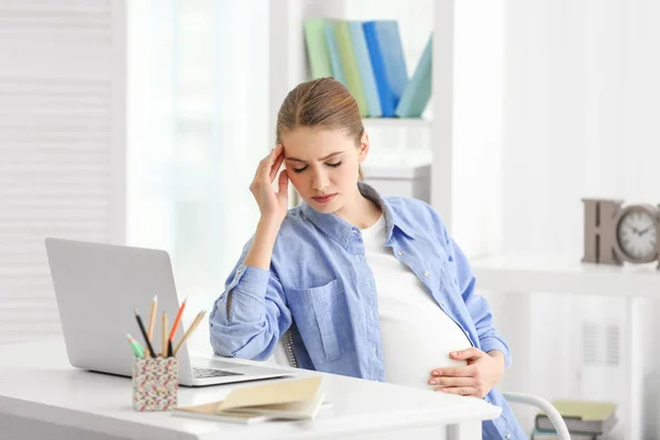 Беременная женщина, страдающая головными болями, сидя на рабочем месте в светлом помещении — стоковое фото