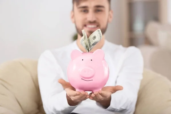Glücklicher junger Mann im Sessel mit Sparschwein und Geld — Stockfoto
