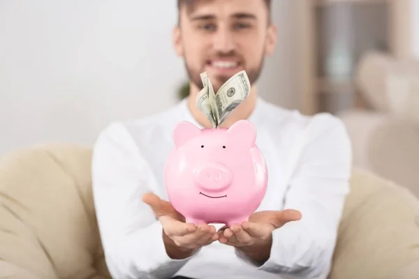 Jovem feliz sentado em poltrona com banco porquinho e dinheiro — Fotografia de Stock