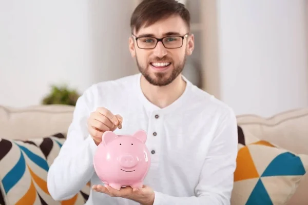 Jovem feliz sentado no sofá e colocando moeda no banco porquinho — Fotografia de Stock