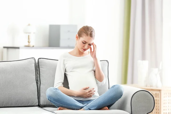 Gravid kvinde, der lider af hovedpine, mens du sidder på sofaen derhjemme - Stock-foto
