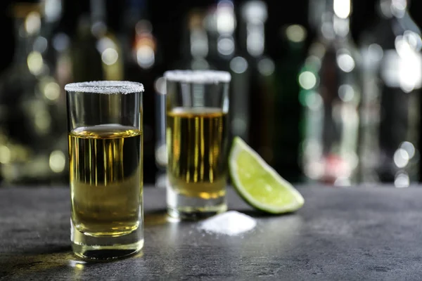 Colpi di tequila dorata con lime succosa e sale su sfondo sfocato di bottiglie di vetro — Foto Stock