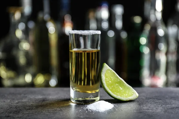 Colpo di tequila dorata con lime succosa e sale su sfondo sfocato di bottiglie di vetro — Foto Stock