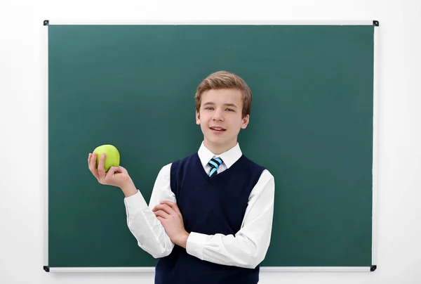 Netter Schüler mit Apfel steht neben Tafel im Klassenzimmer — Stockfoto