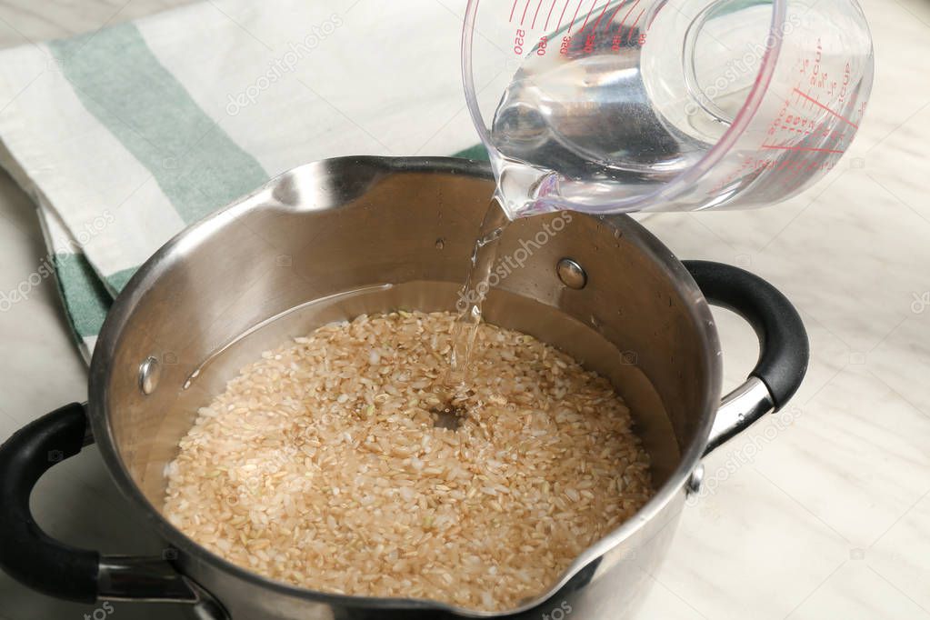 Pouring water into saucepan with brown rice on kitchen table