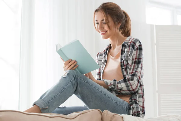 Vacker ung kvinna läser bok nära fönstret hemma — Stockfoto