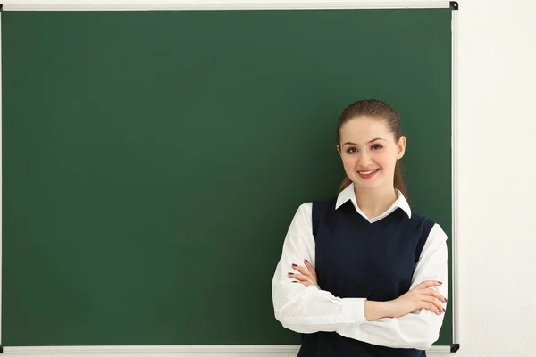 Écolière souriante avec bras croisés debout au tableau noir dans la salle de classe — Photo