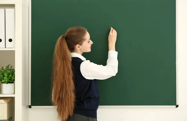 Mooie langharige schoolmeisje schrijven op een schoolbord in de klas — Stockfoto