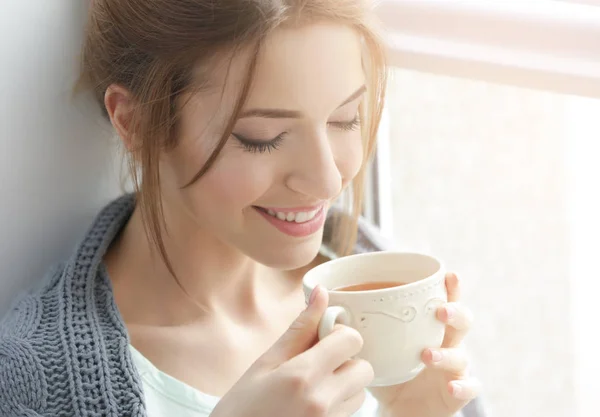 Hermosa joven bebiendo té cerca de la ventana en casa — Foto de Stock