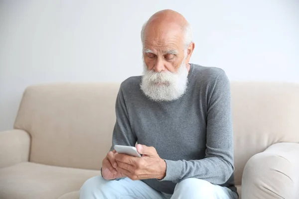 Hombre mayor sentado en el sofá con teléfono inteligente en la habitación — Foto de Stock