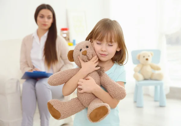 Gadis kecil dengan boneka beruang dan psikolog anak di latar belakang — Stok Foto