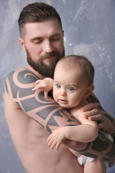Όμορφος τατουάζ νεαρός άνδρας κρατά χαριτωμένο μωράκι σε γκρίζο φόντο — Φωτογραφία Αρχείου