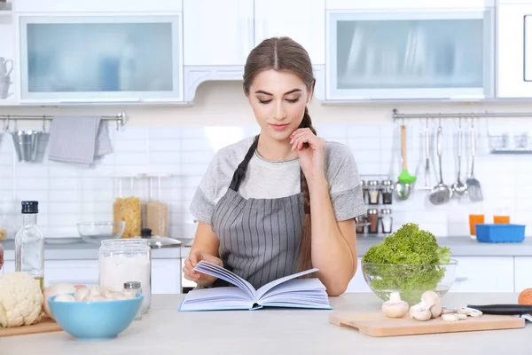 Młoda kobieta z książki kucharskiej przy stole w kuchni — Zdjęcie stockowe