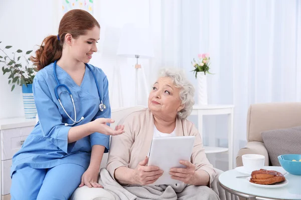 Nurse helping elderly woman to work on tablet pc in light room — Stock Photo, Image