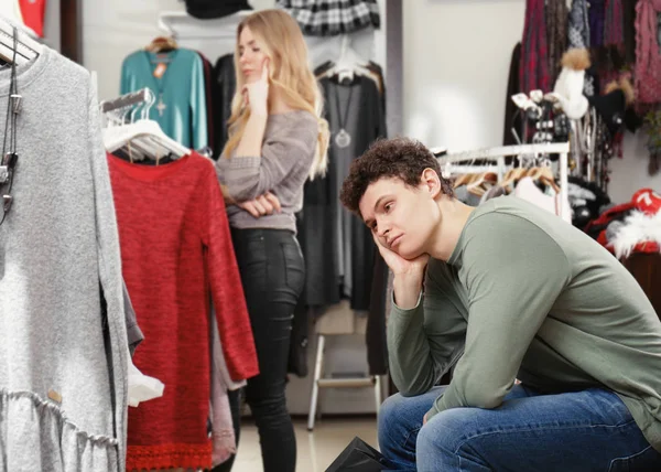 Homem esperando sua namorada enquanto ela escolhe roupas na loja moderna — Fotografia de Stock