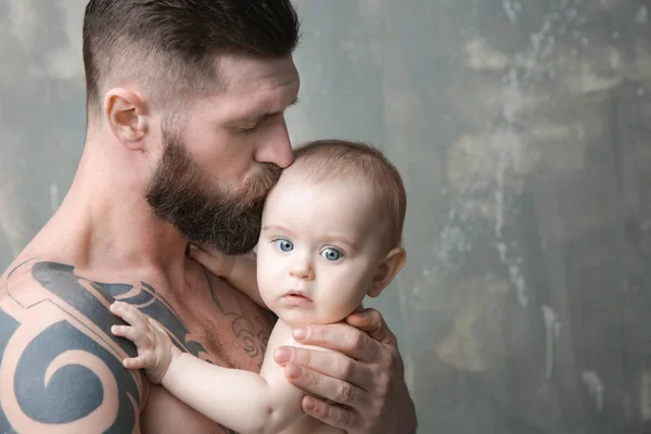 Bonito tatuado jovem segurando bonito pequeno bebê no fundo cinza — Fotografia de Stock