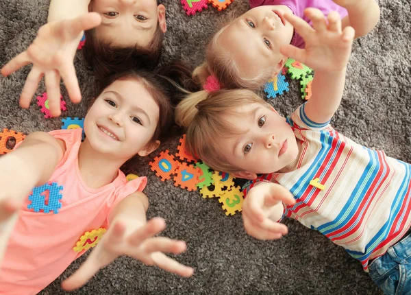 Lindos niños pequeños jugando con figuras coloridas mientras están acostados en la alfombra en casa —  Fotos de Stock