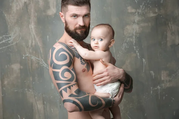 Beau jeune homme tatoué tenant mignon petit bébé sur fond gris — Photo